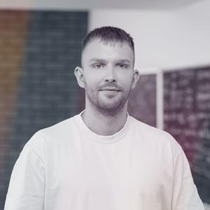 Muž s krátkými vlasy a upraveným plnovousem v bílém tričku, stojí v interiéru s mírným úsměvem; v pozadí je rozmazaná tmavá stěna a tabule s nečitelným textem. Je to Juraj Kruželák, Head of Brand v GymBeam, který vystoupí na konferenci Eshopista: Na vlně influencer marketingu dne 17.6.2025.