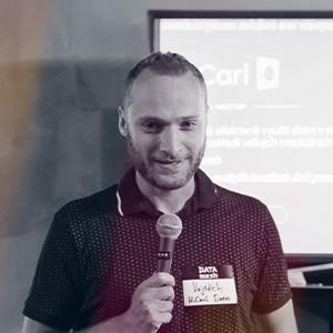 Muž s krátkými vlasy a vousy drží mikrofon a usmívá se při prezentaci. Na sobě má tmavé polo triko s drobným vzorem a jmenovku s nápisem 'DATA meetup – Vojtěch'. V pozadí je rozmazaná prezentace s logem Carl Data. Jedná se o Vojtěcha Matouška, analytika a spoluzakladatele společnosti Carl Data Company, který vystoupí na konferenci Eshopista 17. června 2025.