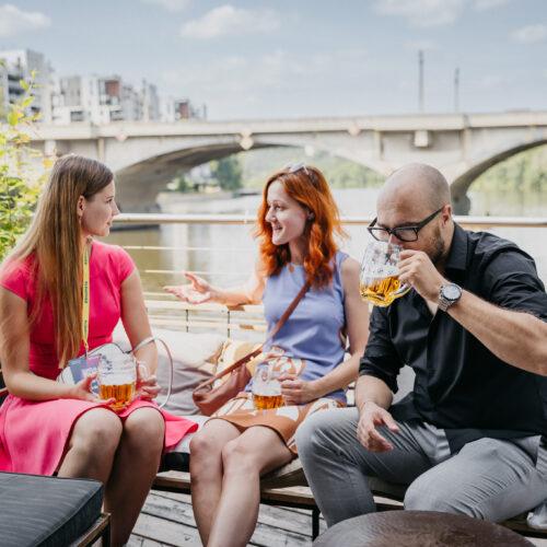 Tři účastníci s lanyardem „ESHOPISTA“ popíjejí pivo na terase u řeky.