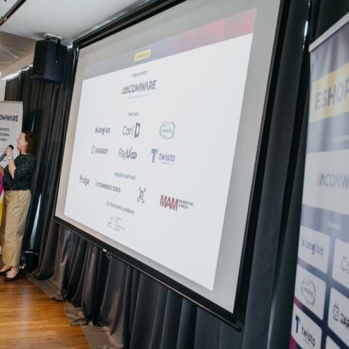 Přednášející na akci ESHOPISTA hovoří vedle projekce s logy ACOMWARE a partnerů.