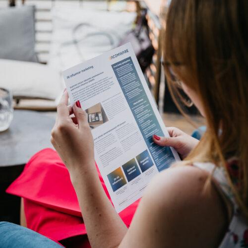 Žena s červenými nehty čte leták ACOMWARE „PR influencer marketing“ v pohodlném venkovním křesle.
